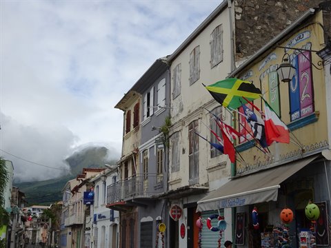 Flags flutter in front of colonial facades beneath cloud-covered Mount Pelée