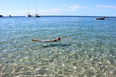 Woman floating in clear turquoise water, sailboats anchored on the horizon
