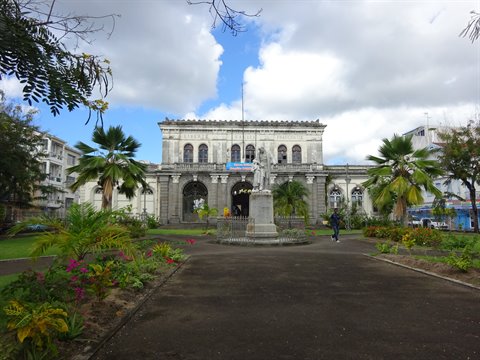 Statue on pedestal before a colonial stone building, among palms and flowered gardens