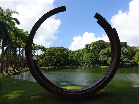 Rusted metal circular sculpture beside a pond surrounded by palm trees
