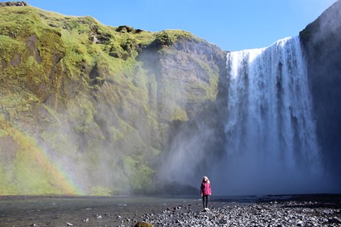Donna in giacca rosa davanti alla cascata di Skogafoss, spruzzi e arcobaleno