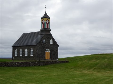 Chiesa in pietra nera con campanile colorato, sul pr verde sotto nuvole grigie, Hvalsnes