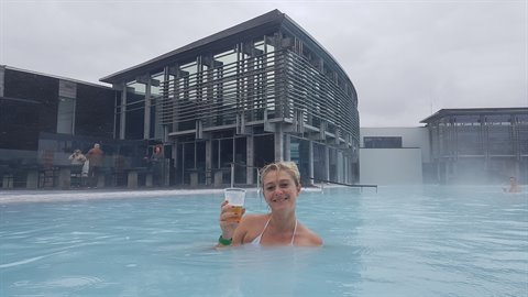 Blue Lagoon: donna sorridente nell'acqua termale con un drink, edificio moderno sullo sfondo