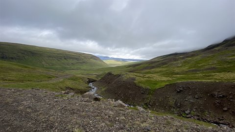Valle verde tra colline muschiate, torrente e cielo grigio, Islanda