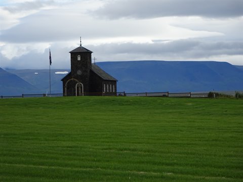 Chiesa nera in pietra su prato verde, montagne blu sullo sfondo, Hvalsnes