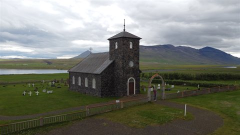 Chiesa di pietra nera con campanile, cimitero con croci bianche, monti e lago