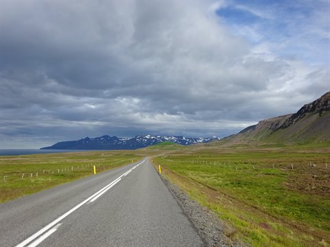 Strada asfaltata tra praterie verdi verso montagne innevate, nuvole dense