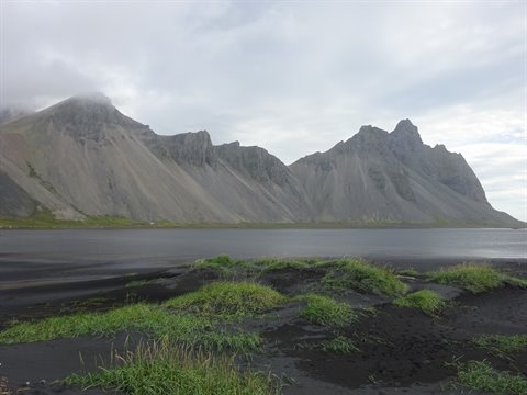 Vestrahorn si specchia nell'acqua sotto nuvole basse, sabbia nera ed erba verde