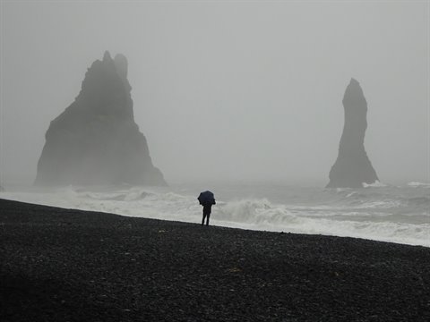 Vík: persona con ombrello sulla spiaggia nera, faraglioni nella nebbia