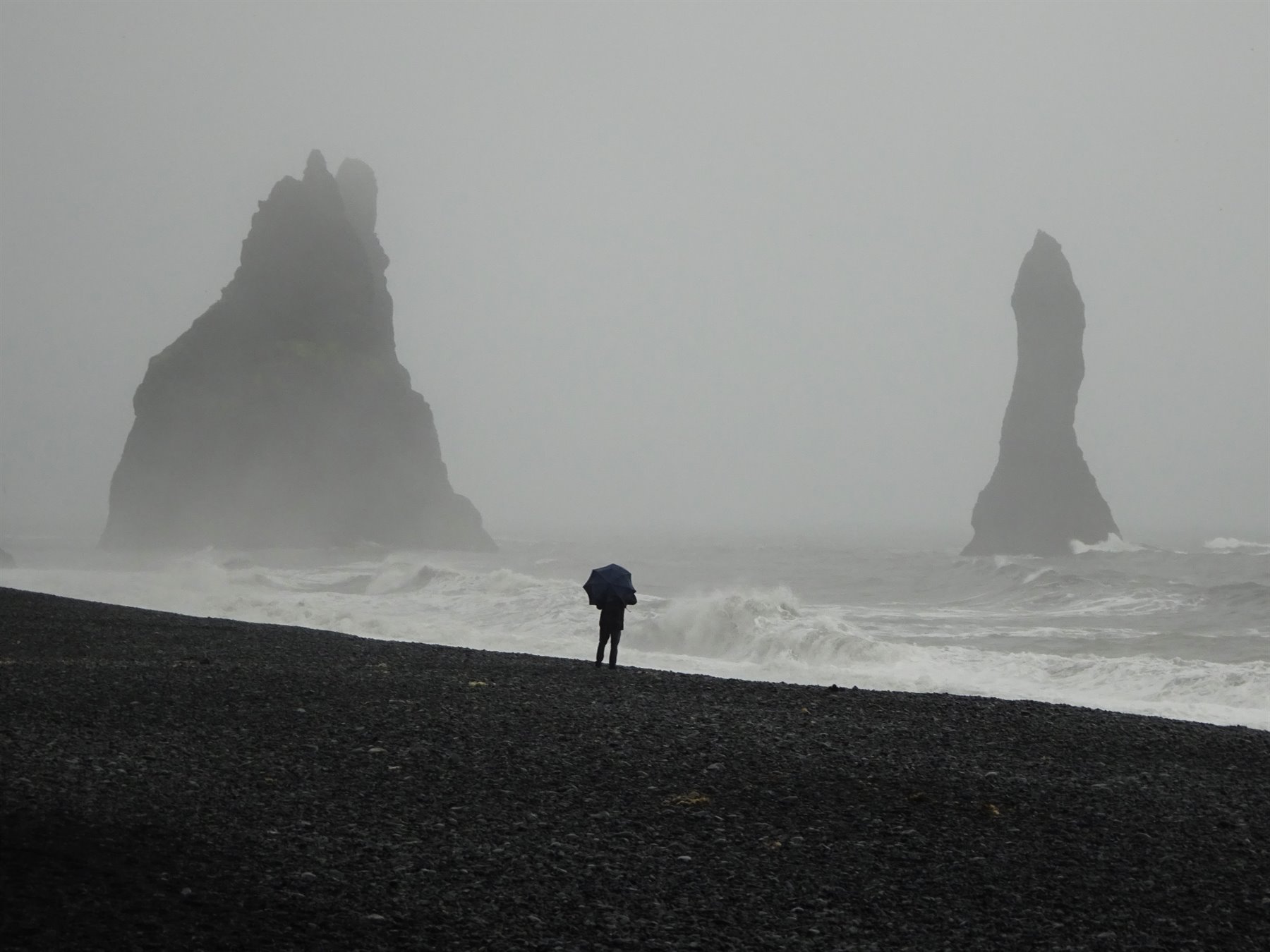 Vík: persona con ombrello sulla spiaggia nera, faraglioni nella nebbia