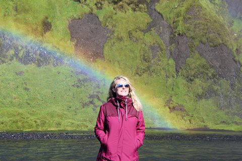 Donna con giacca fucsia davanti alla cascata Skógafoss, arcobaleno sulla roccia muschiosa