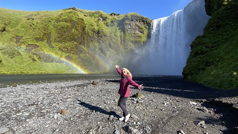 Donna in giacca bordeaux salta gioiosa sulla spiaggia nera sotto la cascata con arcobaleno, Skógafos