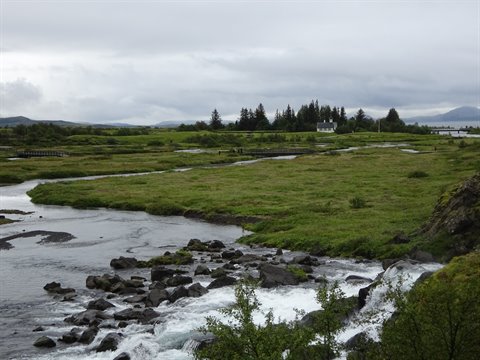 Fiume tra rocce e praterie verdi, case lontane sotto cielo grigioso, Þingvellir