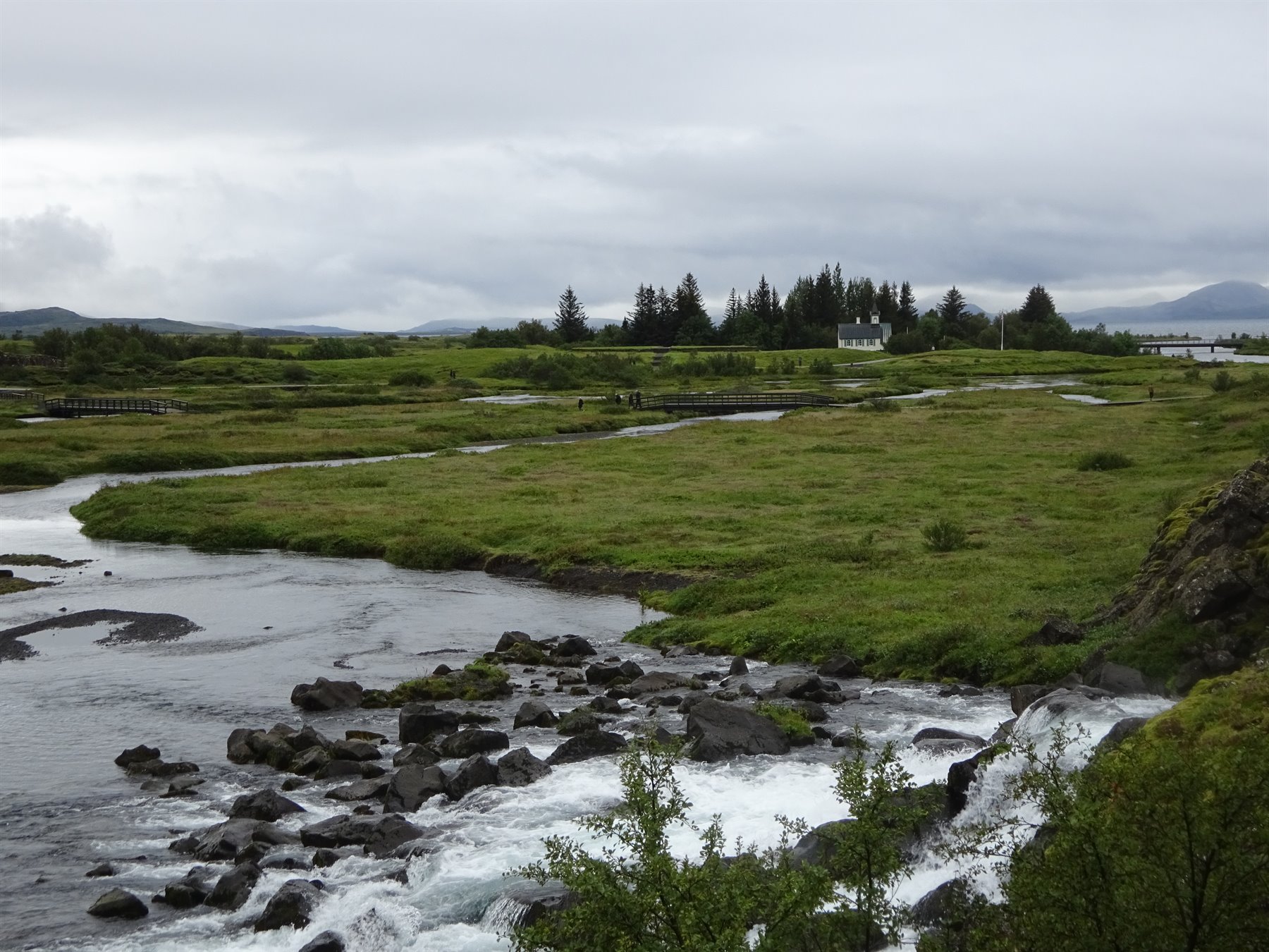 Fiume tra rocce e praterie verdi, case lontane sotto cielo grigioso, Þingvellir