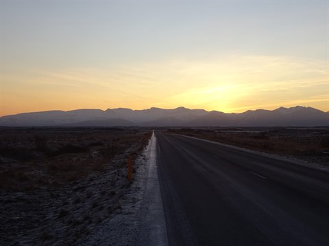 Paved road toward snowy mountains, low golden light on the horizon