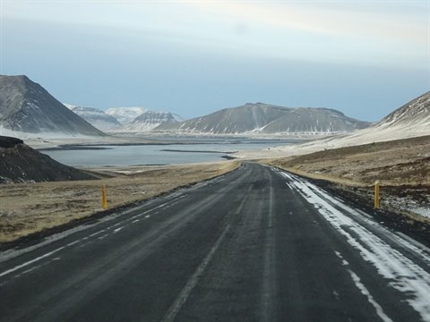 Asphalt road with snow patches toward a fjord, snowy mountains, soft light, travel journal