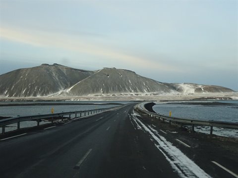 Road with snow leading to snowy volcanic mountains, water on both sides, gray sky, cold light