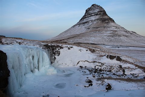Snowy conical mountain and frozen waterfall under pale blue sky