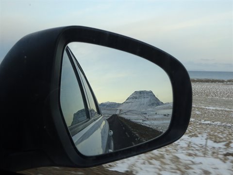 Car side mirror reflecting snowy mountain and road, icy countryside under pale light