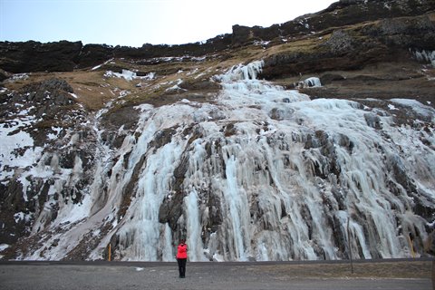 Person in red jacket facing frozen waterfall on rocky cliff, diffused light