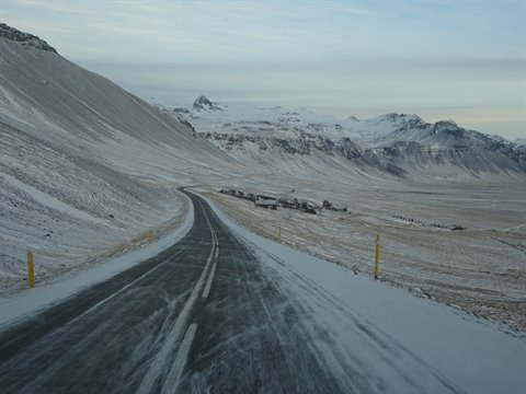 Snowy paved road winding between mountains under grey sky, cold light, road trip