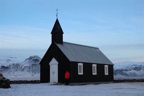 Black wooden church in snow, person in red jacket by the door, cold light