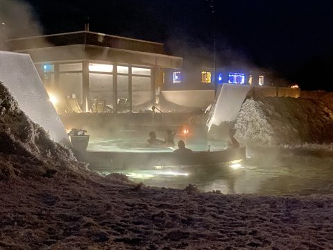 Bathers in steaming hot pool amid snow, illuminated building at night