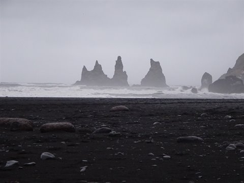 Snowy rock formations emerge from fog above a wide black beach, soft diffused light