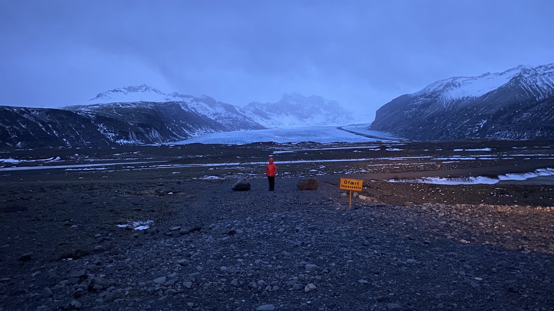 Traveler in red jacket facing a glacier between snowy mountains, twilight light
