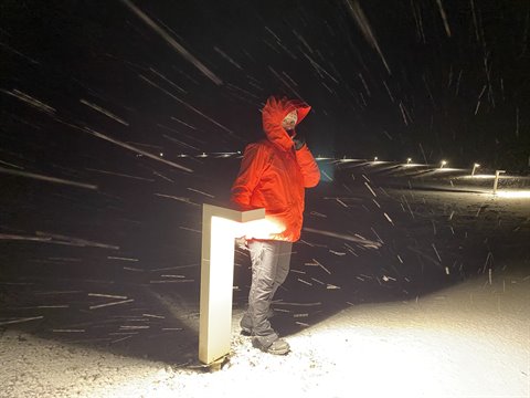 Person in orange jacket beside glowing marker in falling snow at night