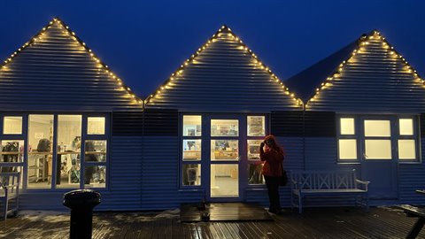 Woman photographing outside white wooden beach huts with string lights at blue hour
