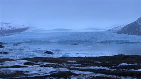 Blue glacier surrounded by snowy mountains reflected in frozen lake under diffuse light