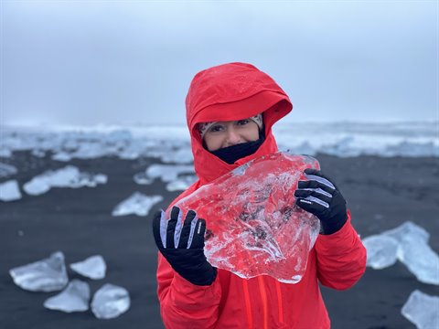 Traveler in red jacket holding an ice chunk on black sand beach, soft light