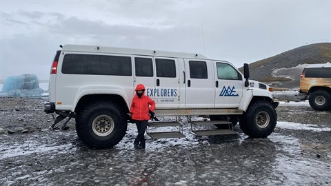 Traveler in red jacket beside a large white off-road truck on snowy ground, overcast sky