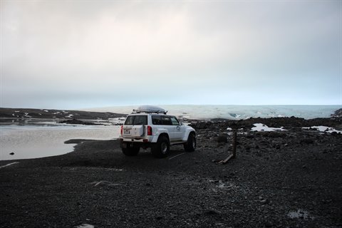 White off-road vehicle on black volcanic sand in front of a glacier, cloudy sky