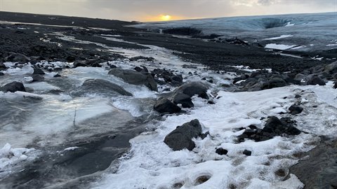 Frozen stream over black volcanic rocks before a glacier, low light