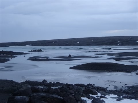 Frozen lake with dark islands among black rocks and snow, cold cloudy light