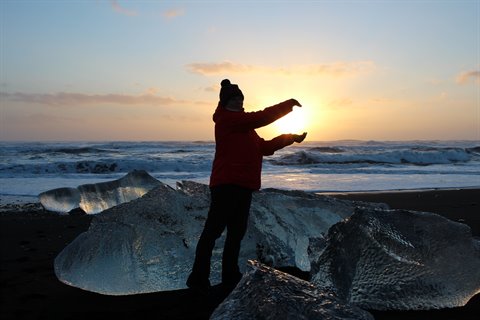 Person in red jacket frames the sun between ice chunks on black sand beach