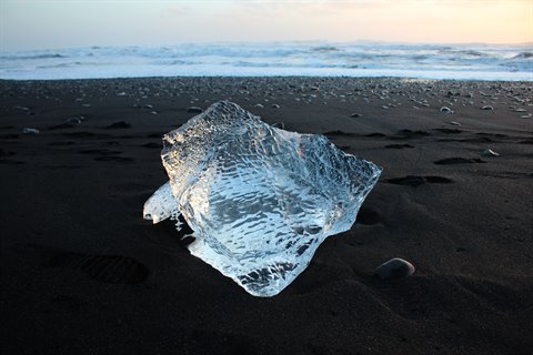 Clear ice block on black sand, waves and golden light behind