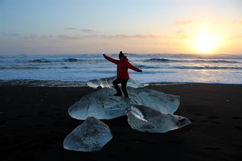 Woman in red jacket on ice blocks, black beach, golden light