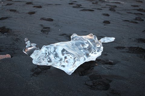 Transparent ice chunk on black volcanic sand, footprints around, cold light