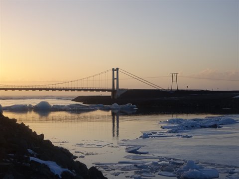 Concrete suspension bridge over icy river, reflections on calm water, golden light