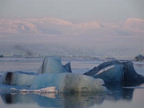 Large blue icebergs float on calm water, snowy mountains fading in the background