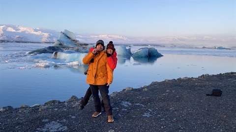 Woman in orange jacket kisses man in beanie on black sand beach, floating icebergs, golden light