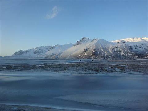 Snow-covered mountains under blue sky, icy plain in foreground, cold morning light