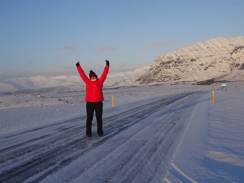 Traveler in red jacket raising arms on snowy road between snow-capped mountains