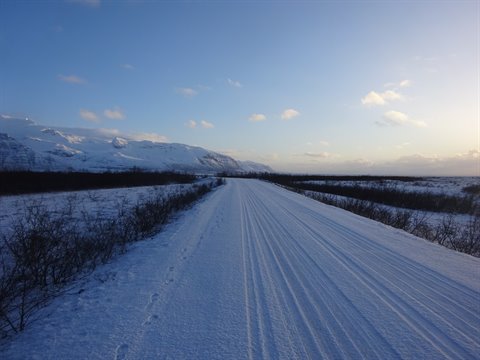 Snow-covered road crossed on foot between bushes and mountains, cold morning light