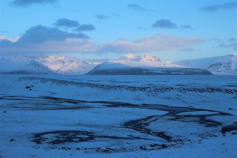 Snowy mountains glowing under blue morning light, frozen plain with dark cracks in foreground