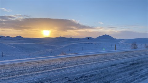 Snowy road among frozen hills, low sun through clouds, cold light