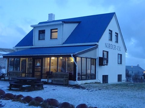 White house with blue roof, Nordur-Vik sign, cold light on snow and wooden benches